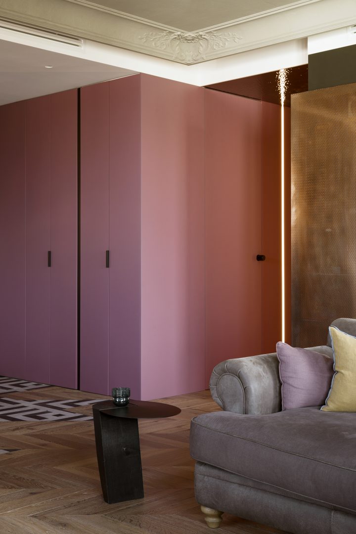 Szara sofa, czarny stolik i różowa zabudowa szaf ze sztukaterią na suficie